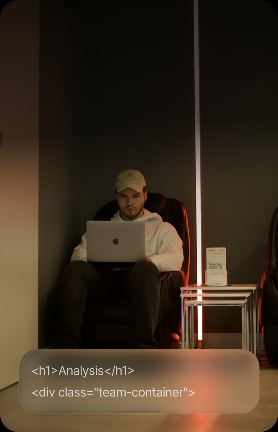 A person wearing a baseball cap and hoodie sits in a chair, focused on a laptop.