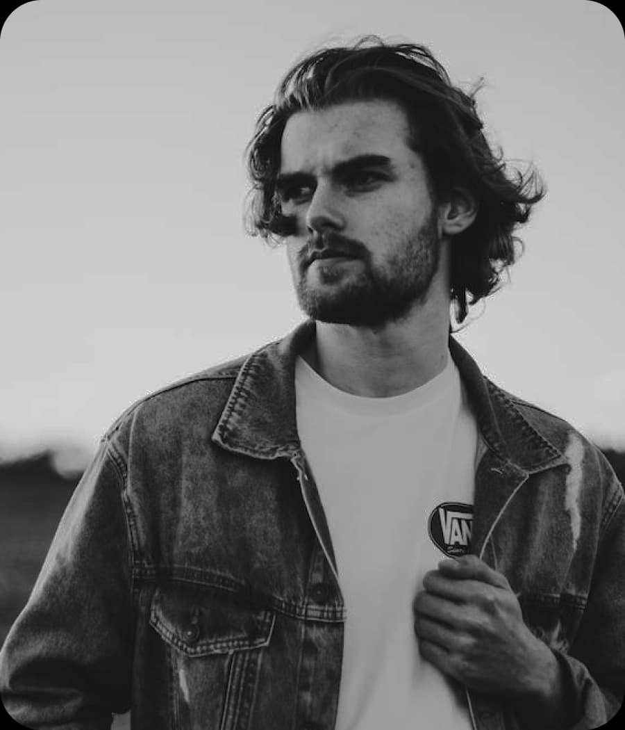 Man with long hair wearing a denim jacket and t-shirt looking to the side.