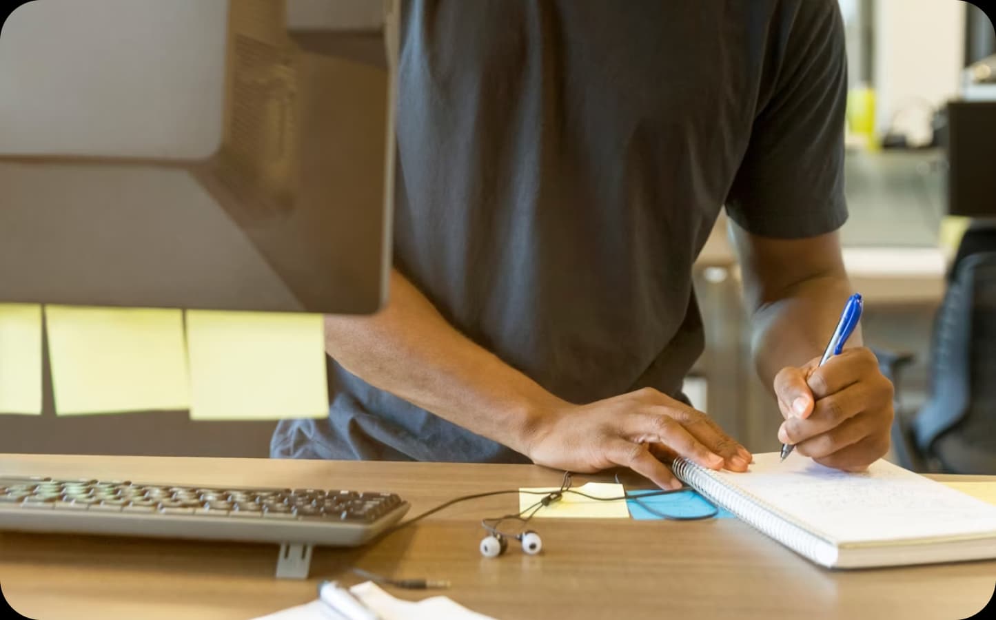 Person's hands writing in a notebook at a desk with a computer.