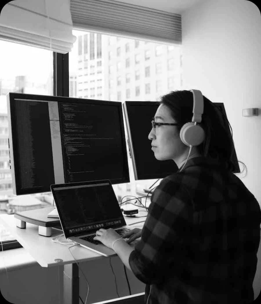A woman wearing headphones and glasses types on a laptop in front of multiple computer monitors displaying code.